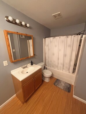 A bathroom with a wooden vanity, a white sink, a toilet, a shower with a white curtain, and a mirror above the vanity. - KC Strong: Beautiful 2 BR Home, Close to stadiums and downtown. (Independence)