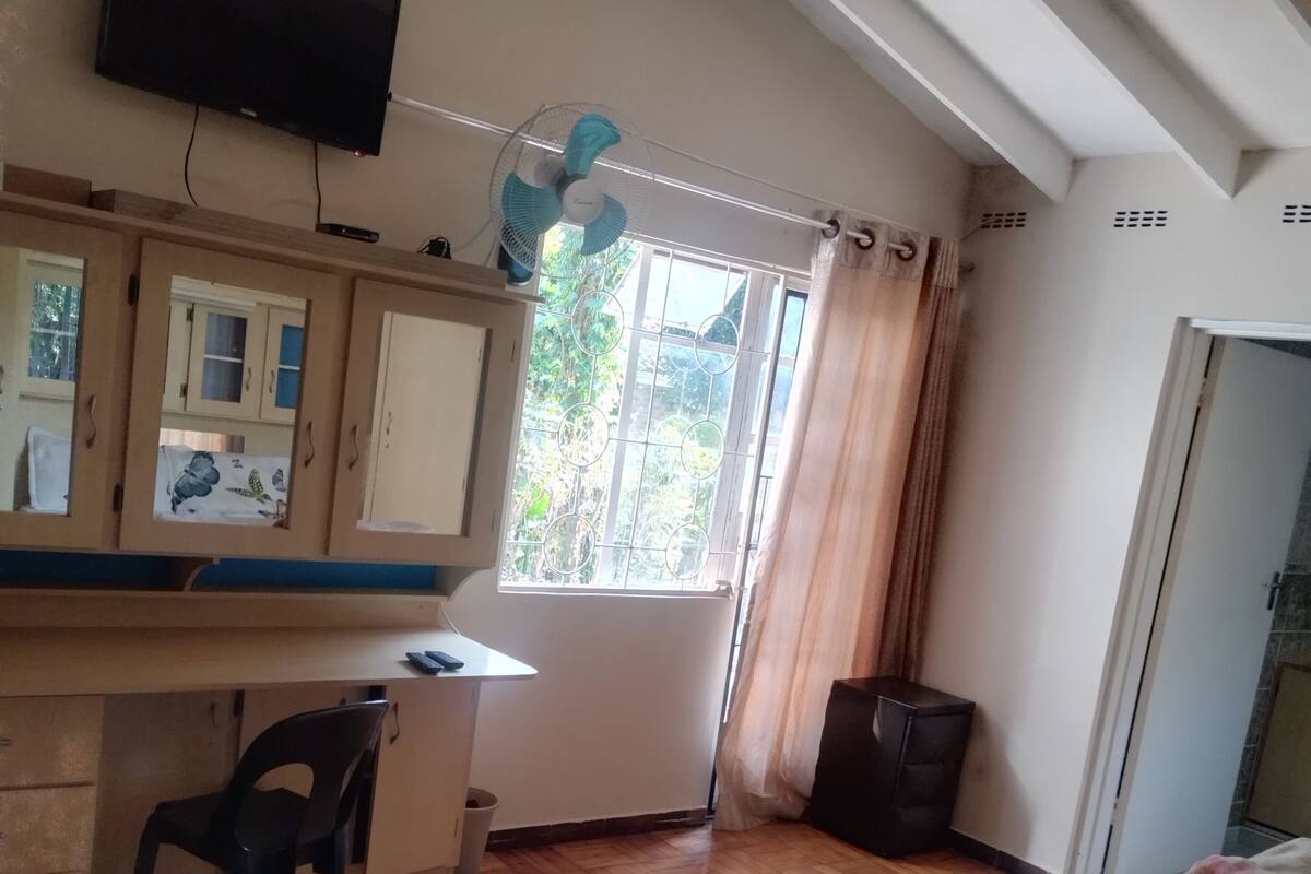 Un dormitorio con cama, escritorio, silla, televisor, ventana con cortinas y ventilador de techo.