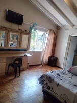 Un dormitorio con cama, escritorio, silla, televisor, ventana con cortinas y ventilador de techo.
