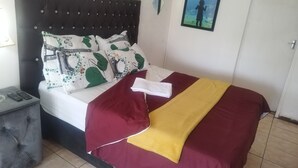 A bed with a dark headboard, white bedding, and decorative pillows. There is a folded towel on the bed, a remote control on a side table, and a framed picture on the wall. - J & J Paradise Guesthouse  (Pietermaritzburg)