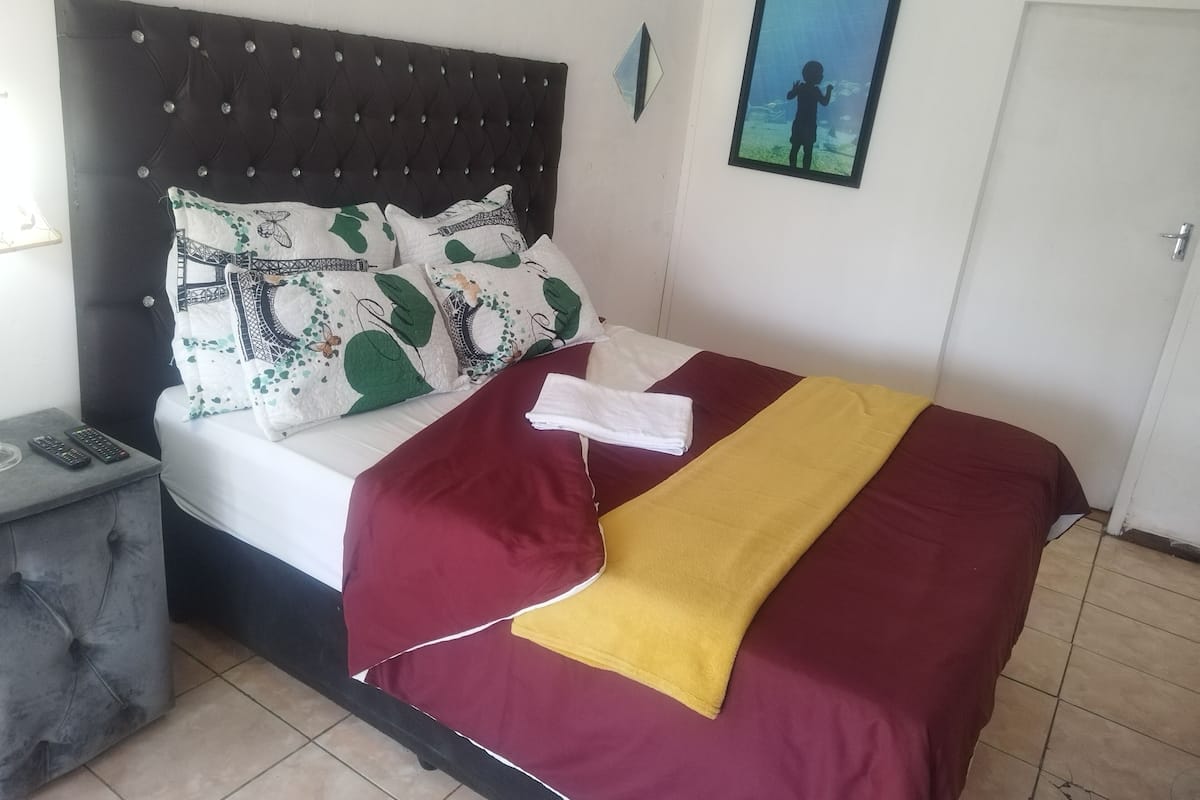 Una cama con cabecera oscura, ropa de cama blanca y almohadas decorativas. Hay una toalla doblada sobre la cama, un control remoto en una mesita de noche y un cuadro enmarcado en la pared.