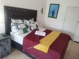 Una cama con cabecera oscura, ropa de cama blanca y almohadas decorativas. Hay una toalla doblada sobre la cama, un control remoto en una mesita de noche y un cuadro enmarcado en la pared.