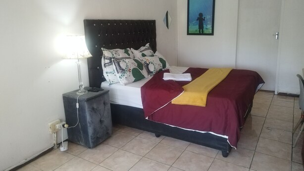 A bedroom with a bed, bedside table, lamp, and a painting on the wall. - J & J Paradise Guesthouse  (Pietermaritzburg)