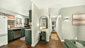 Een moderne keuken met donkergroene kastjes, een vaatwasser en een magnetron. Een gang met vloerkleed leidt naar een woonkamer met een groene bank.