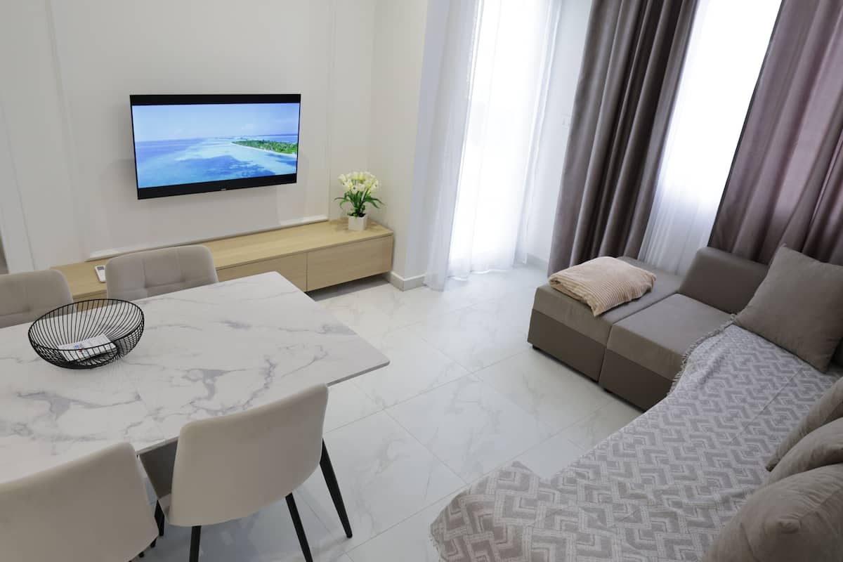 A modern living room with a flat-screen TV, a dining area with a marble table, and a sofa with a patterned throw.