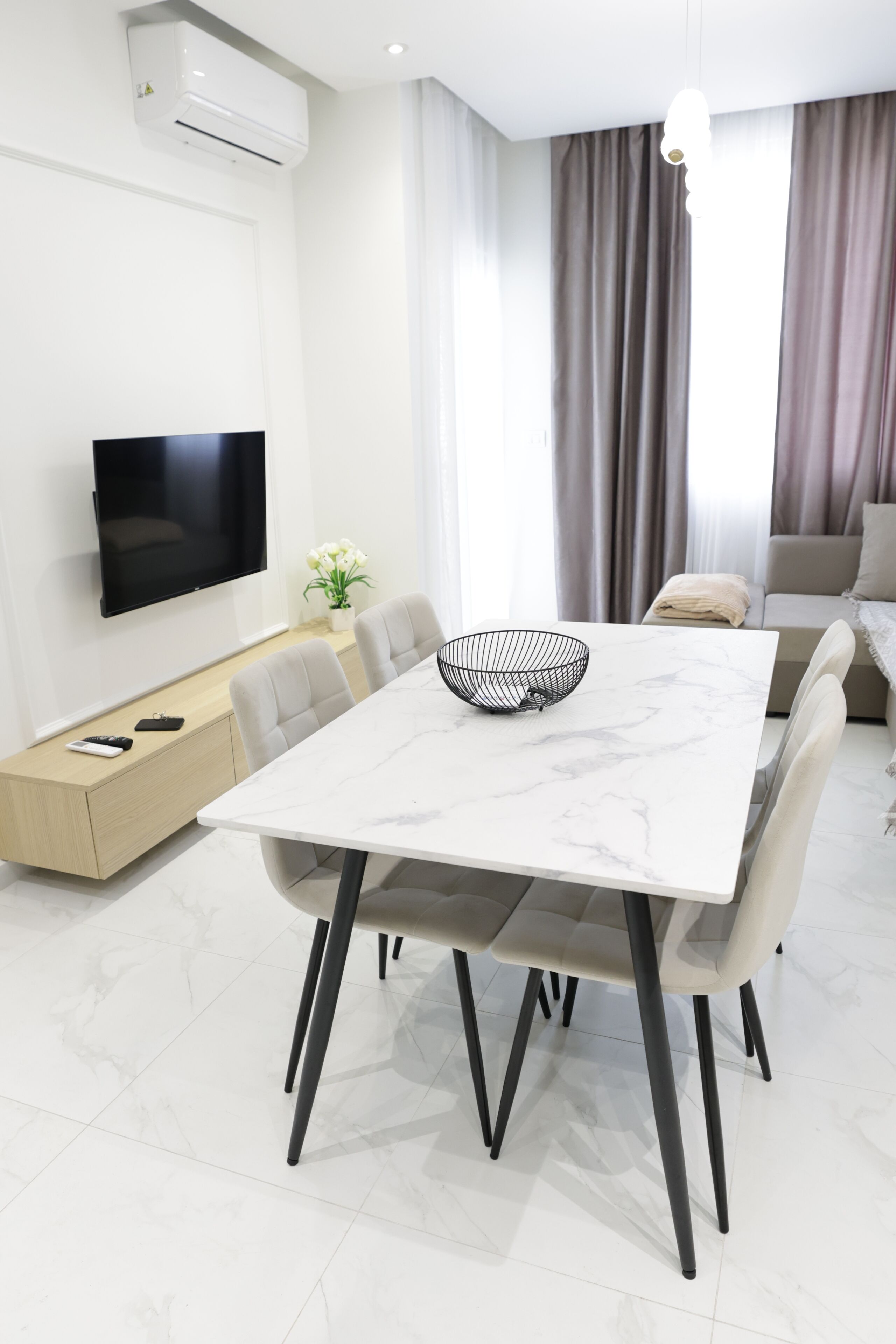 A modern living room with a flat-screen TV, a dining table with chairs, a sofa, and a window with curtains.