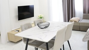 Un salon moderne doté d’un téléviseur à écran plat, d’une table à manger avec des chaises, d’un canapé et d’une grande fenêtre agrémentée de rideaux.