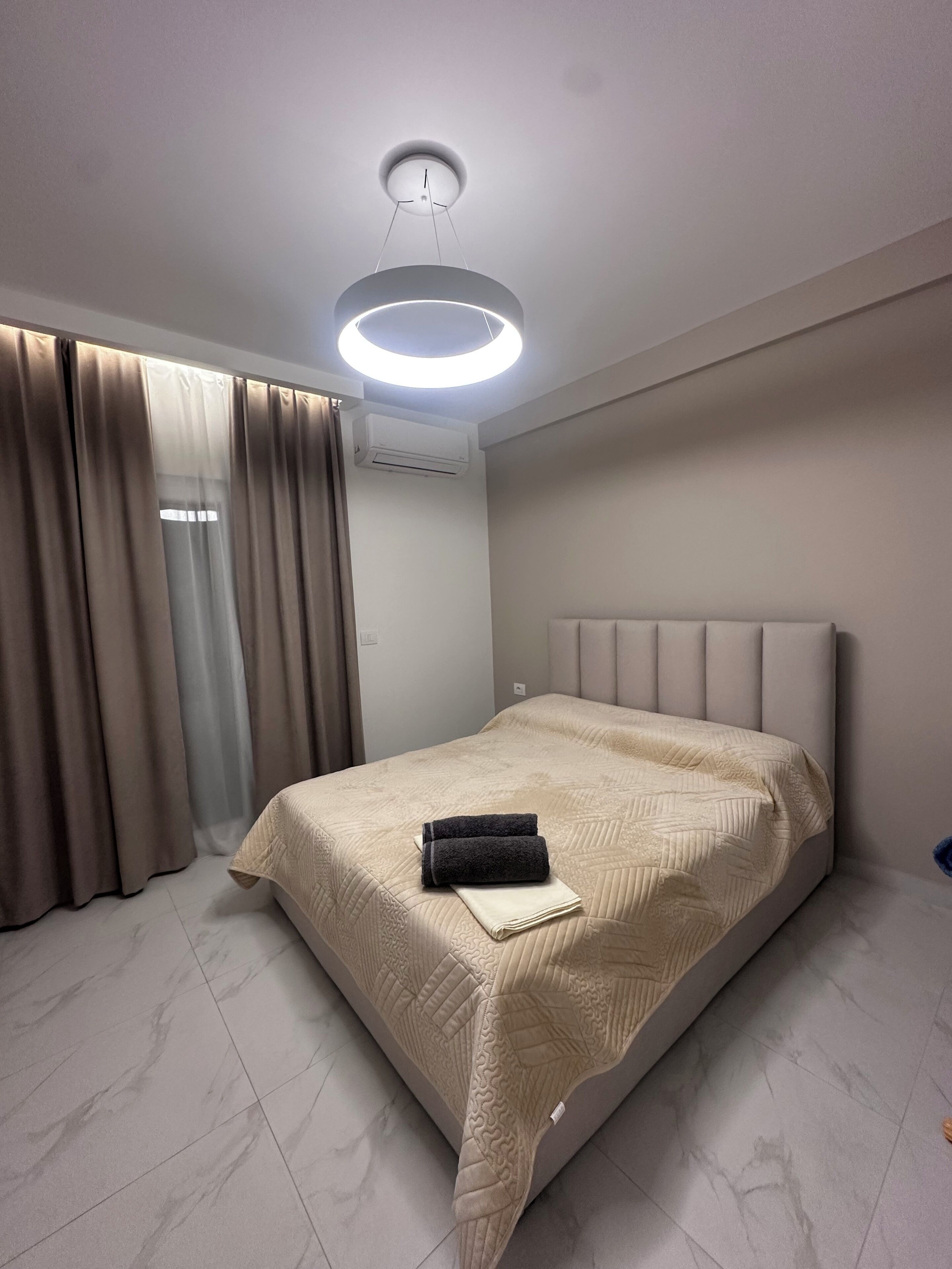 A modern hotel room with a bed, curtains, an air conditioning unit, and a ceiling light fixture.