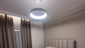A modern hotel room with a bed, curtains, an air conditioning unit, and a ceiling light fixture.