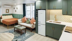 A compact room with a bed, sofa, small table, kitchenette, and a refrigerator.