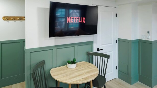 A room with a TV mounted on the wall displaying 'NETFLIX,' a round wooden table with chairs, and a small plant on the table.