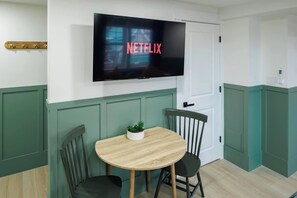 Et værelse med et fjernsyn monteret på væggen, der viser 'NETFLIX', et rundt træbord med stole og en lille plante på bordet.