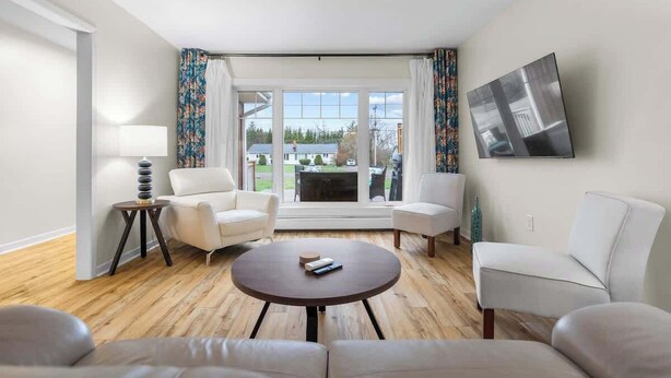A modern living room with a round coffee table, two white armchairs, a sofa, and a large window with curtains. - Beach Bliss: Charming 3-Bedroom Home (Brackley)