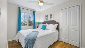 A bedroom with a bed, a ceiling fan, a window with curtains, and a door. - Beach Bliss: Charming 3-Bedroom Home (Brackley)