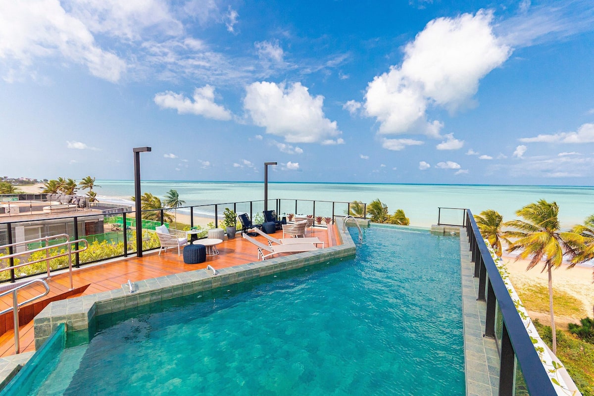 Ein Pool mit Blick auf den Ozean, Sitzgelegenheiten im Freien und Palmen.
