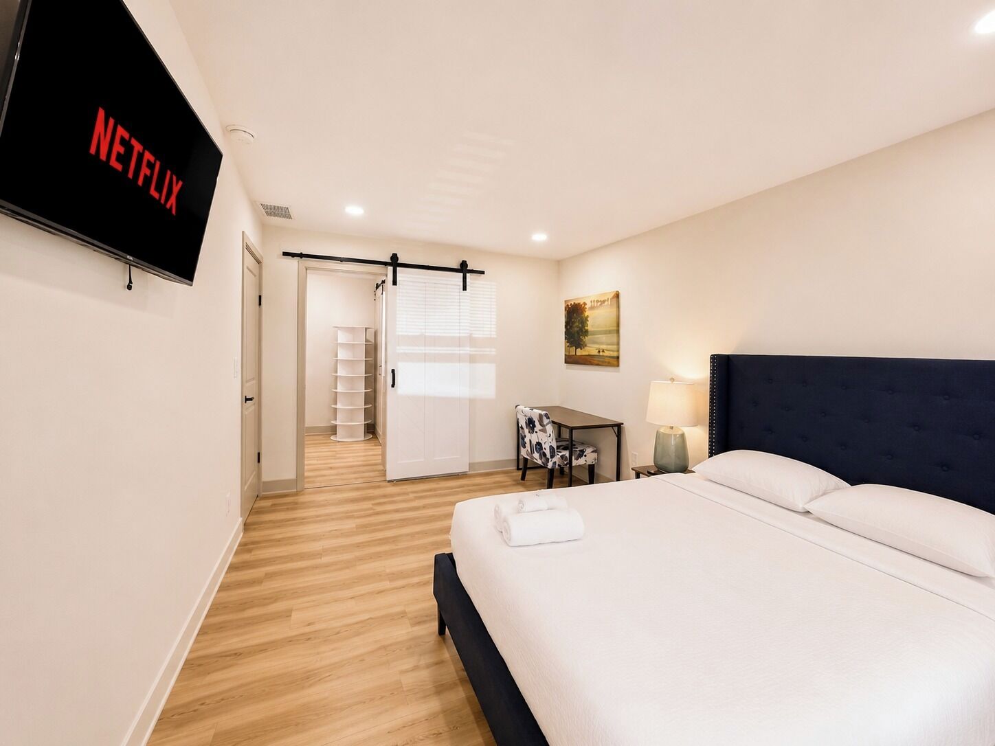 Una habitación de hotel con cama, escritorio, silla, televisor mostrando 'NETFLIX' y puerta que da a un pasillo.