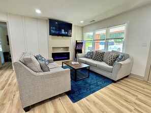 A living room with two sofas, a coffee table, a flat-screen TV, a fireplace, and a window with blinds. - NEW Brick Ranch Near Braves & Six Flags (Mableton)