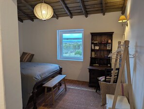 A bedroom with a bed, a small bench, a wooden dresser, a window with a view, and a hanging light fixture.