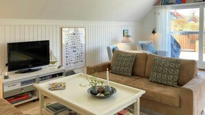 Ein Wohnzimmer mit Fernseher, einem weißen Couchtisch mit Kerze, einer braunen Couch mit gemusterten Kissen, einem Bücherregal und einem Fenster mit Blick auf ein Haus.