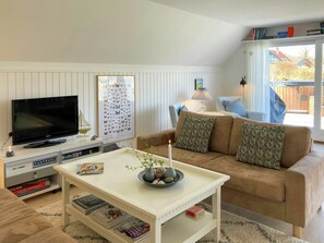 Ein Wohnzimmer mit Fernseher, einem weißen Couchtisch mit Kerze, einer braunen Couch mit gemusterten Kissen, einem Bücherregal und einem Fenster mit Blick auf ein Haus.