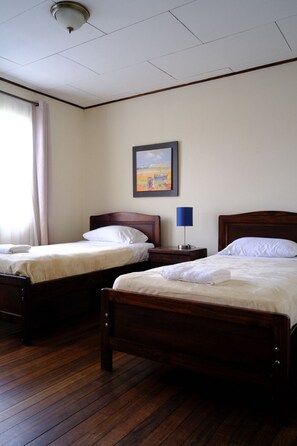 Une chambre d’hôtel avec deux lits en bois, une table de chevet avec une lampe, un tableau encadré au mur et une fenêtre avec des rideaux.