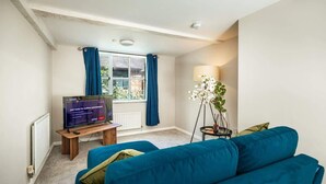 A living room with a blue sofa, a television, a lamp, and a window with curtains. - Central Chester Recently Renovated 1BR Flat (Chester)
