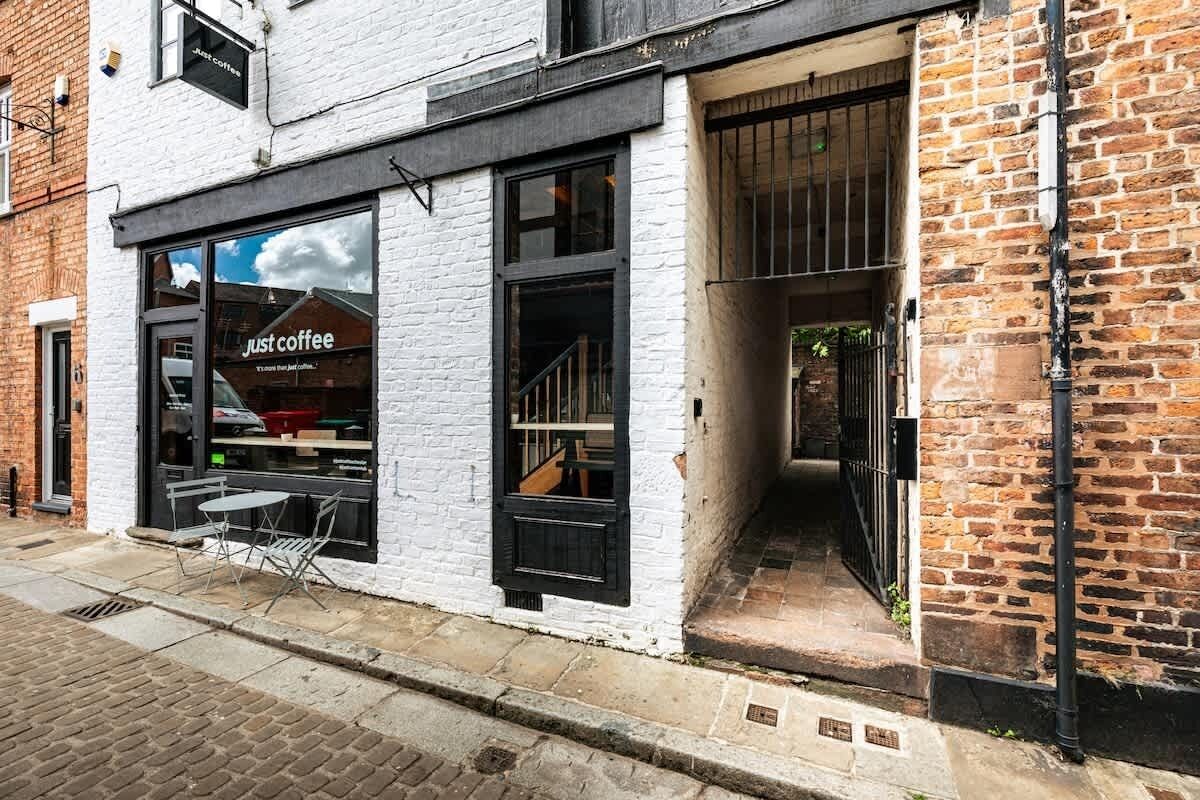 Ein Café mit weißer Fassade, schwarzen Fensterrahmen und einem sichtbaren Außenbereich mit Tischen und Stühlen.