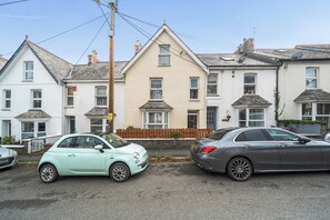Una calle residencial con dos coches aparcados, una casa de fachada blanca y una valla de madera.