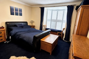 A bedroom with a bed, a wooden bench, a nightstand, a lamp, a wardrobe, and a bay window with curtains. - 1 Bed - Sleeps 4 - Parking - Pets - Town Centre (Wadebridge)