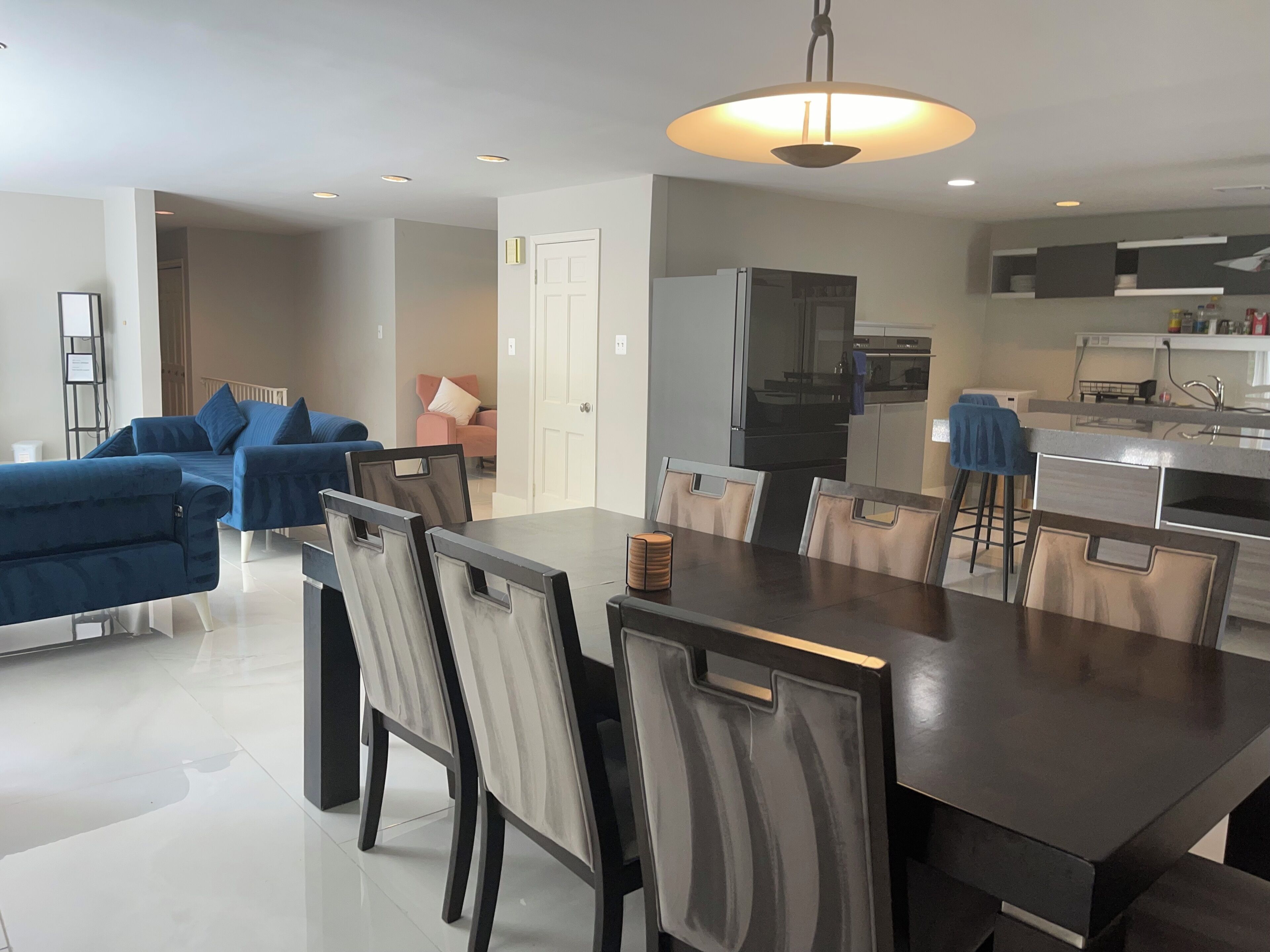A modern living room with a dark wooden dining table and chairs, a blue sofa set, and a kitchen area with a refrigerator and stove.