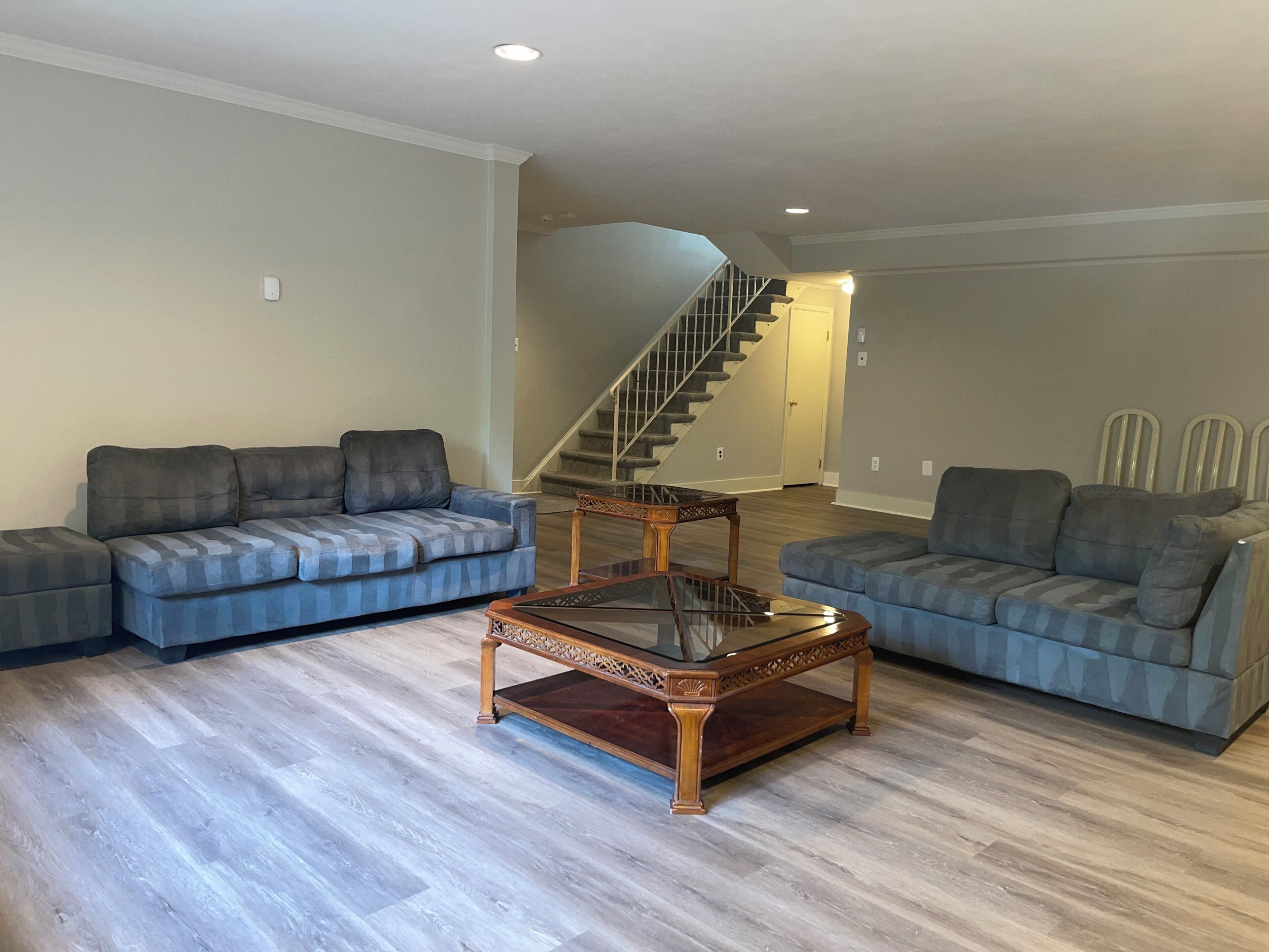 Ein geräumiges Wohnzimmer mit zwei grauen Sofas, einem Glastisch und Holzboden. Im Hintergrund ist eine Treppe zu sehen.