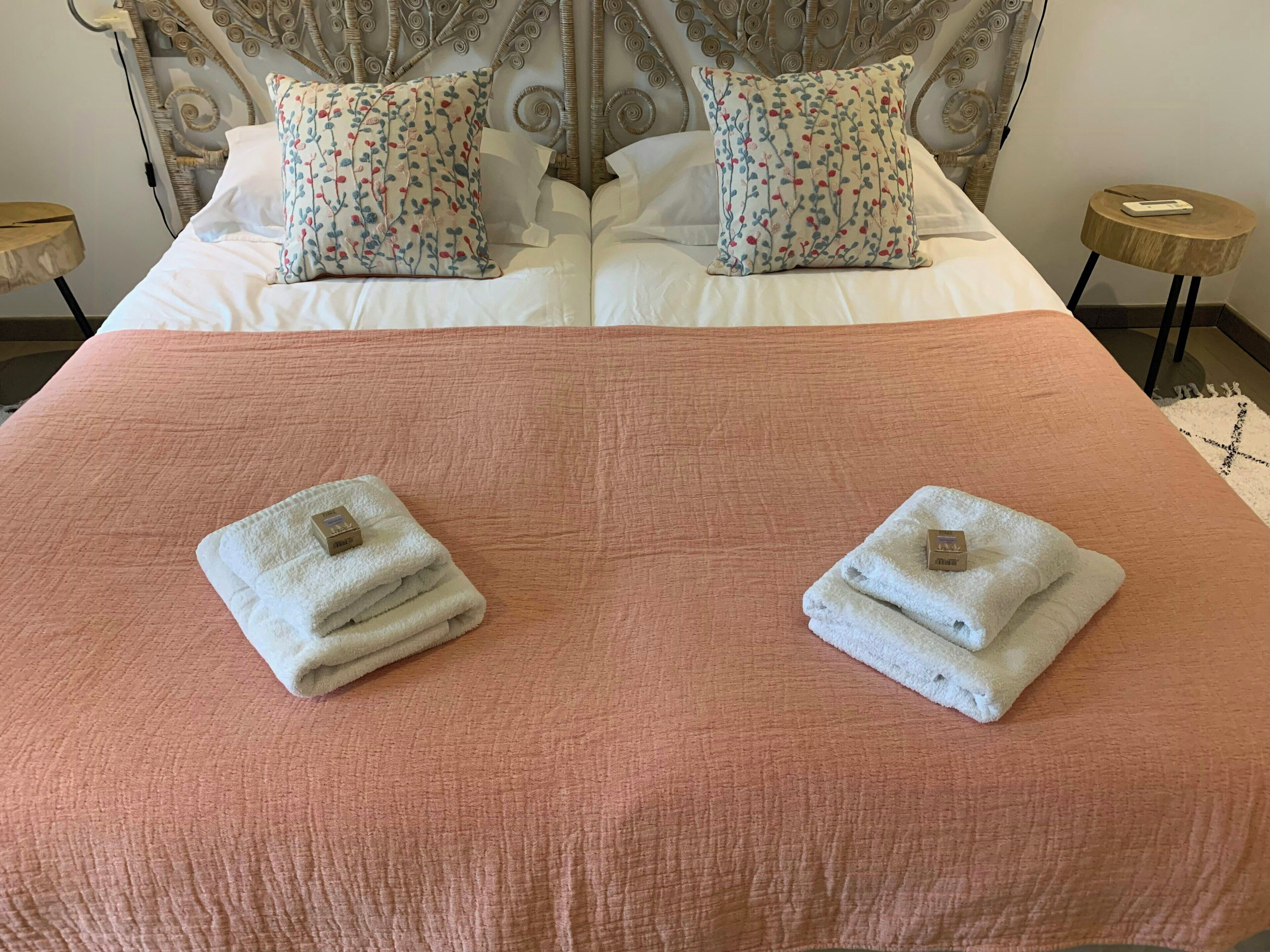 A neatly made bed with a pink bedspread, two pillows with a floral pattern, and two folded towels with a tag on them.