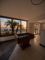 Una mesa de fútbol de mesa en una habitación con ventanas amplias que dan a una playa al atardecer.