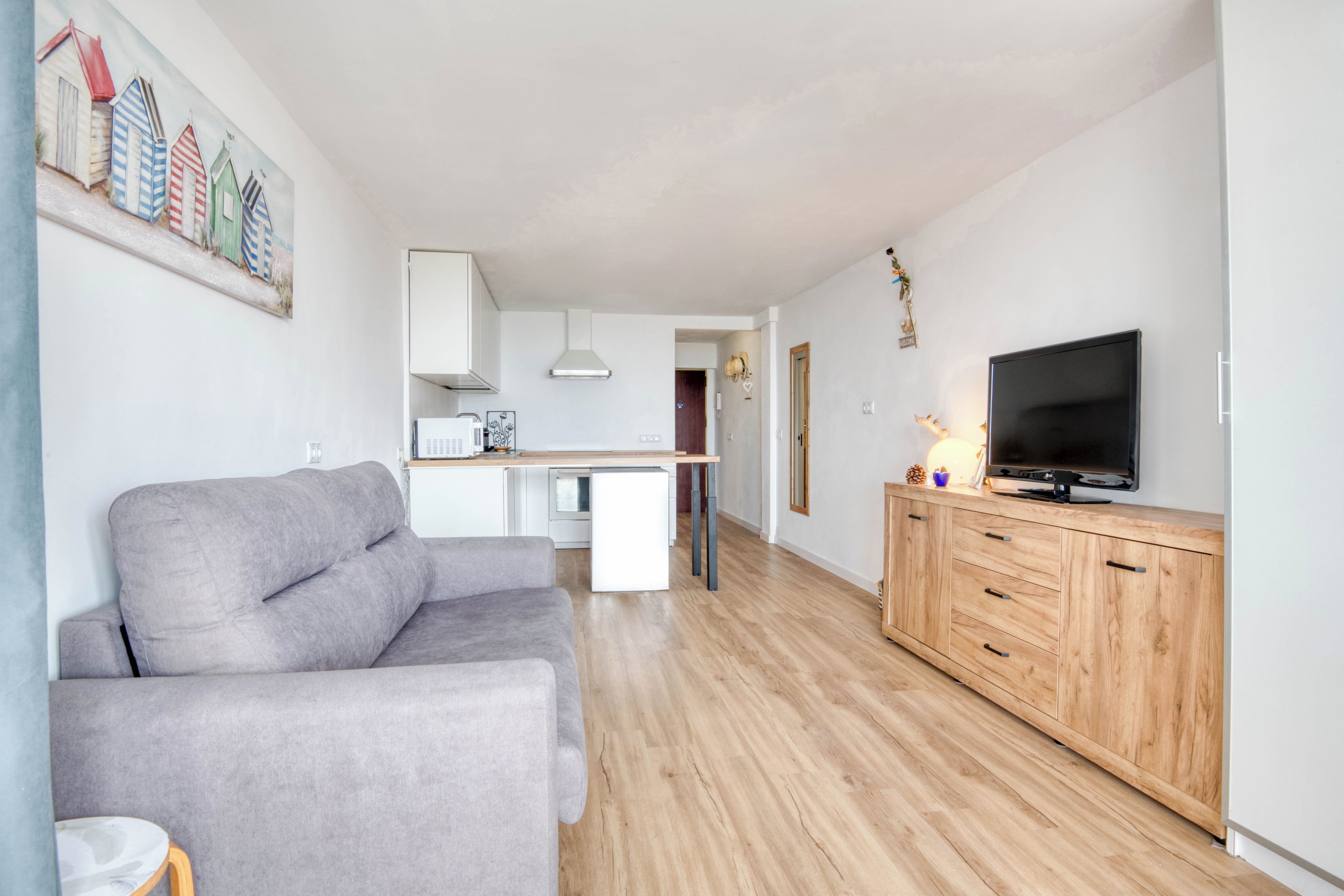 Een moderne woonkamer met een grijze bank, een houten kast met een flatscreen televisie en een keuken op de achtergrond.