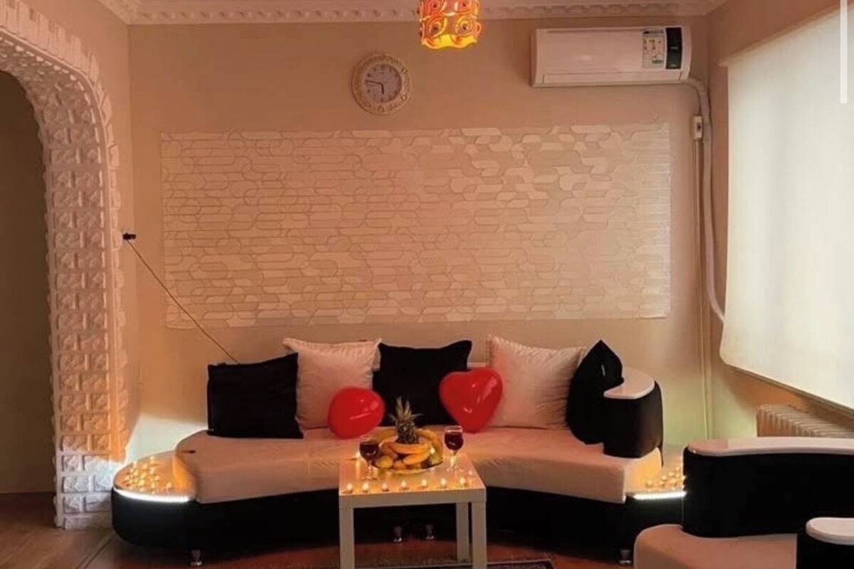 A living room with a white sofa, red heart-shaped pillows, a small table with candles, and a decorative hanging light.