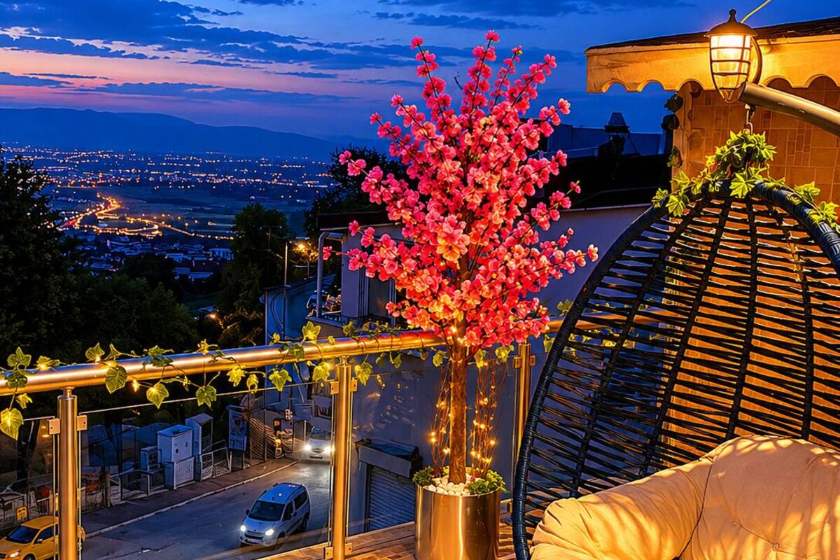 A balcony with a hanging chair, a potted tree, and a view of a cityscape at dusk.