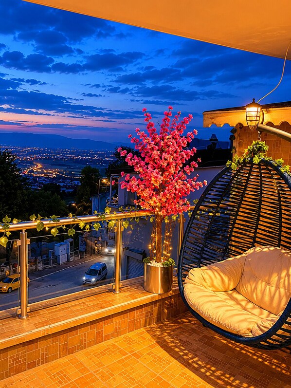 Ein Balkon mit einer Hängematte, einem Topfbaum und Blick auf eine städtische Silhouette in der Abenddämmerung.