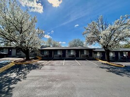 A row of motel rooms with a parking lot in front, trees, and a clear sky.