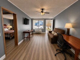 A room with a bed, a desk, a chair, a ceiling fan, a TV, and a window with a view of the outside.