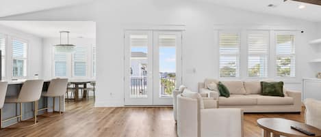 A spacious living area with a wooden floor, a sofa, a dining table with chairs, and large windows with blinds.