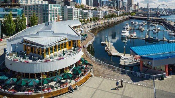 Ein Restaurant am Wasserufer mit Außenbereich, ein Yachthafen mit Booten und eine Stadtansicht im Hintergrund.