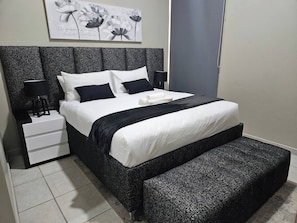 Ein ordentlich eingerichtetes Schlafzimmer mit einem Bett, Nachttischen, einer Bank und einem an der Wand befestigten Kunstwerk.