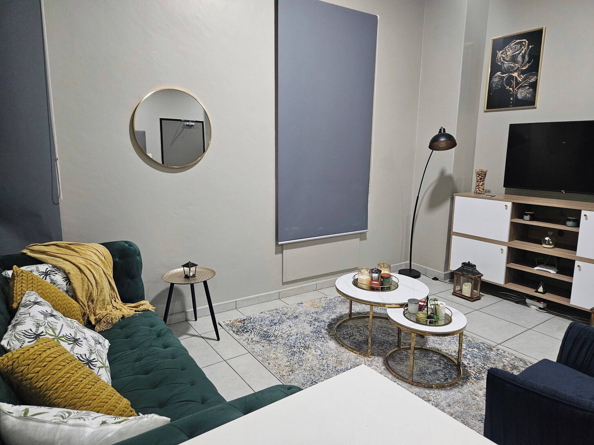 Ein Wohnzimmer mit einem grünen Sofa, einem weißen Couchtisch, einem runden Spiegel, einer Stehlampe und einem Fernseher auf einem Schrank.