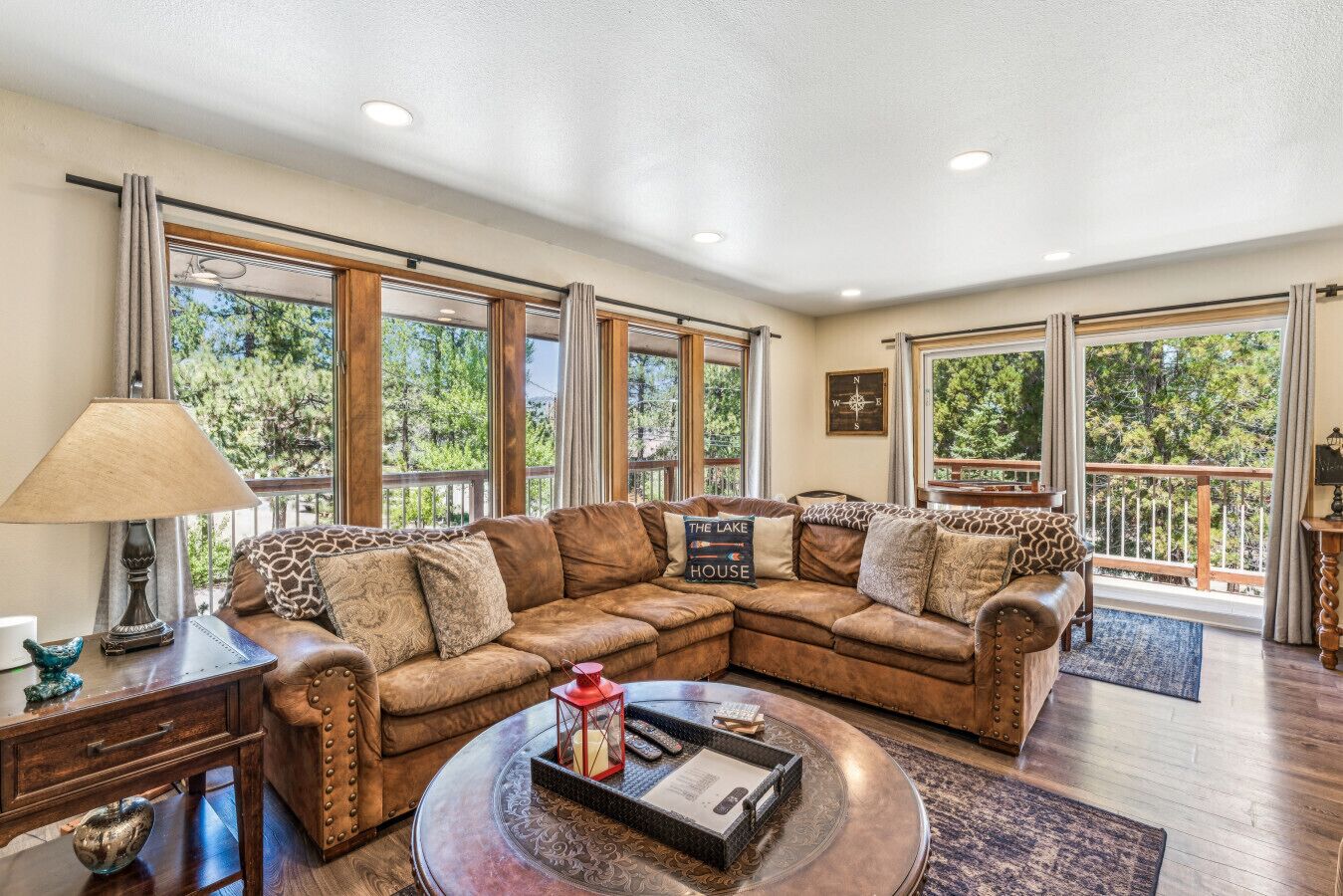 A spacious living room with a large sectional sofa, a round coffee table, and multiple windows offering views of a wooded area.