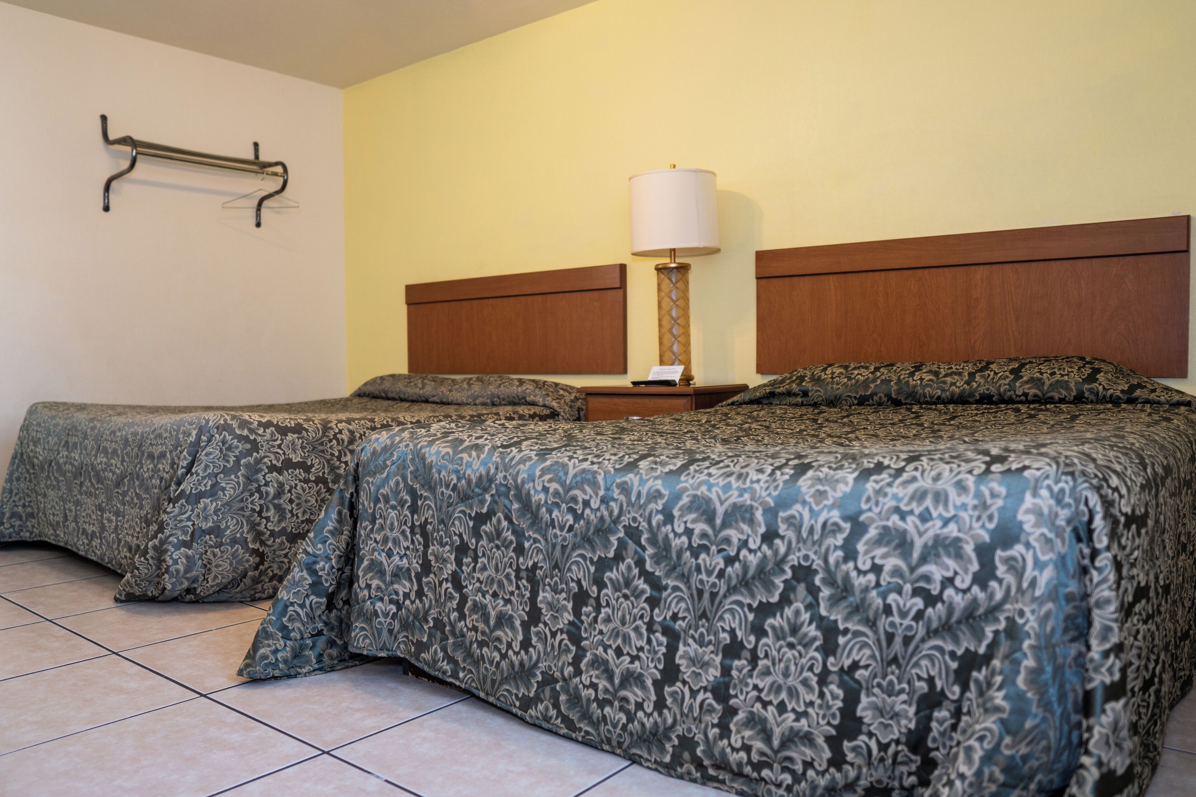 A hotel room with two beds, a bedside table with a lamp, and a wall-mounted towel rack.