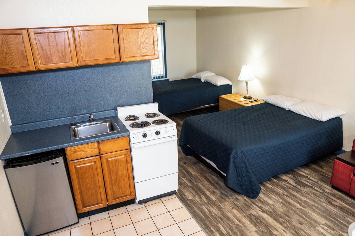 A compact room with a kitchenette, a bed, and a bedside table with a lamp.