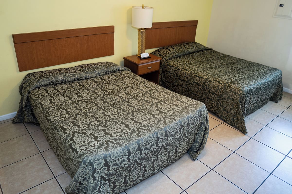 A hotel room with two beds, a nightstand with a lamp, and a plain wall.