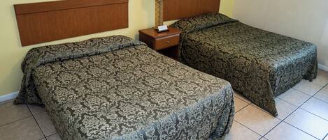 A hotel room with two beds, a nightstand with a lamp, and a plain wall.