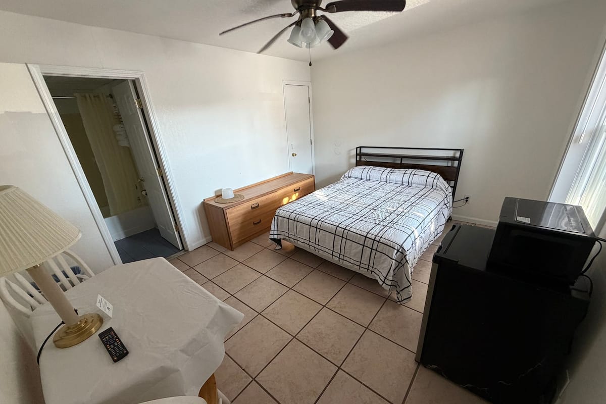 A bedroom with a bed, a desk, a chair, a ceiling fan, a TV, and a window.