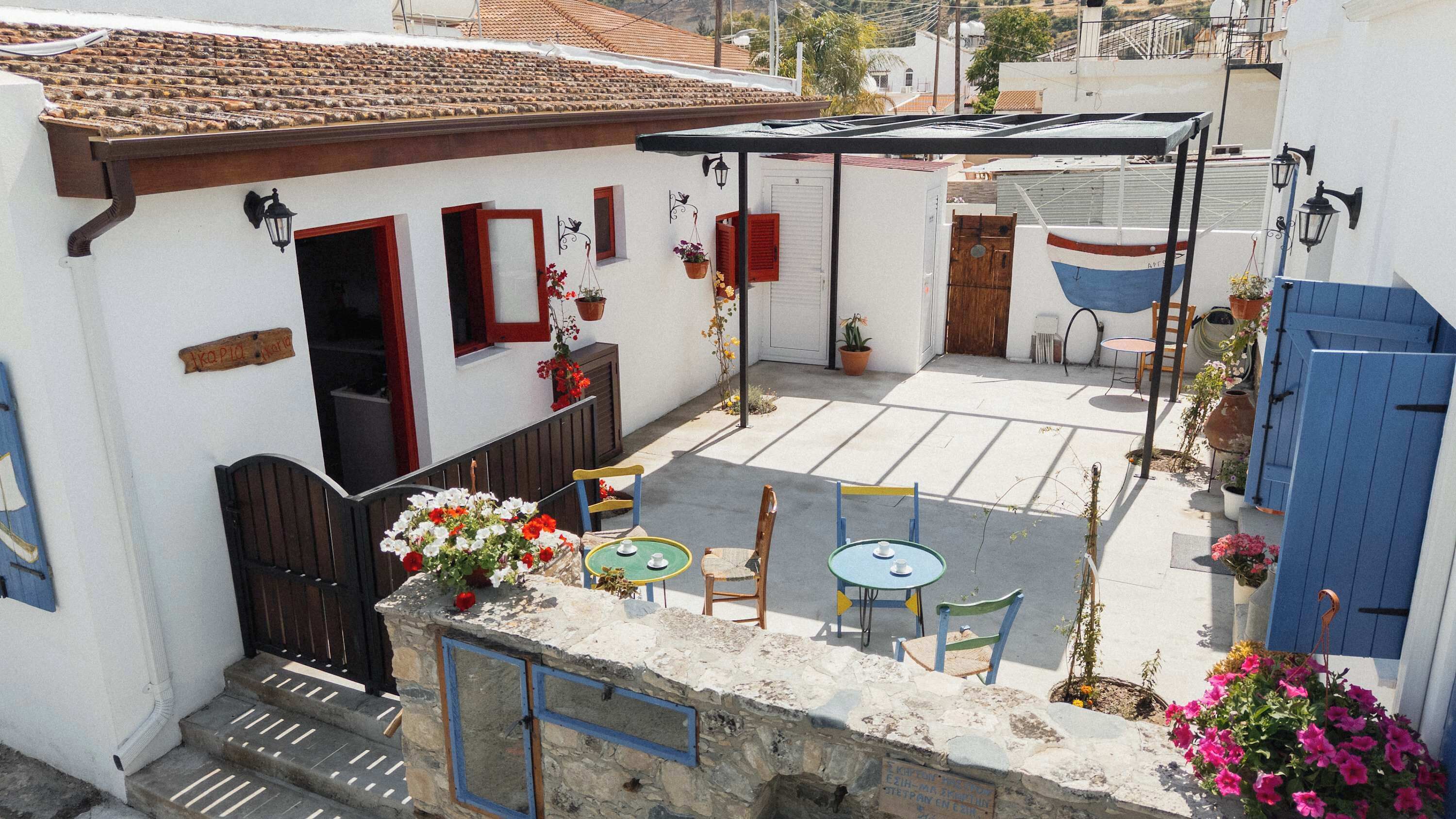 Un patio con un banco de piedra, una mesa y sillas. Hay plantas en macetas y una puerta azul.